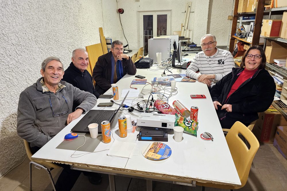 Cinq adhérents du club assis autour de la grande table du local, avec des ordinateurs portables, un vidéoprojecteur et de quoi grignoter